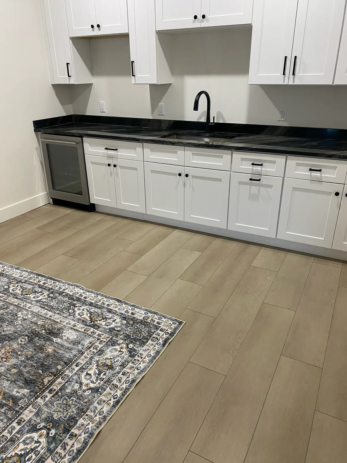 Greige wide-plank vinyl flooring in a butler’s pantry with white shaker cabinets and black granite