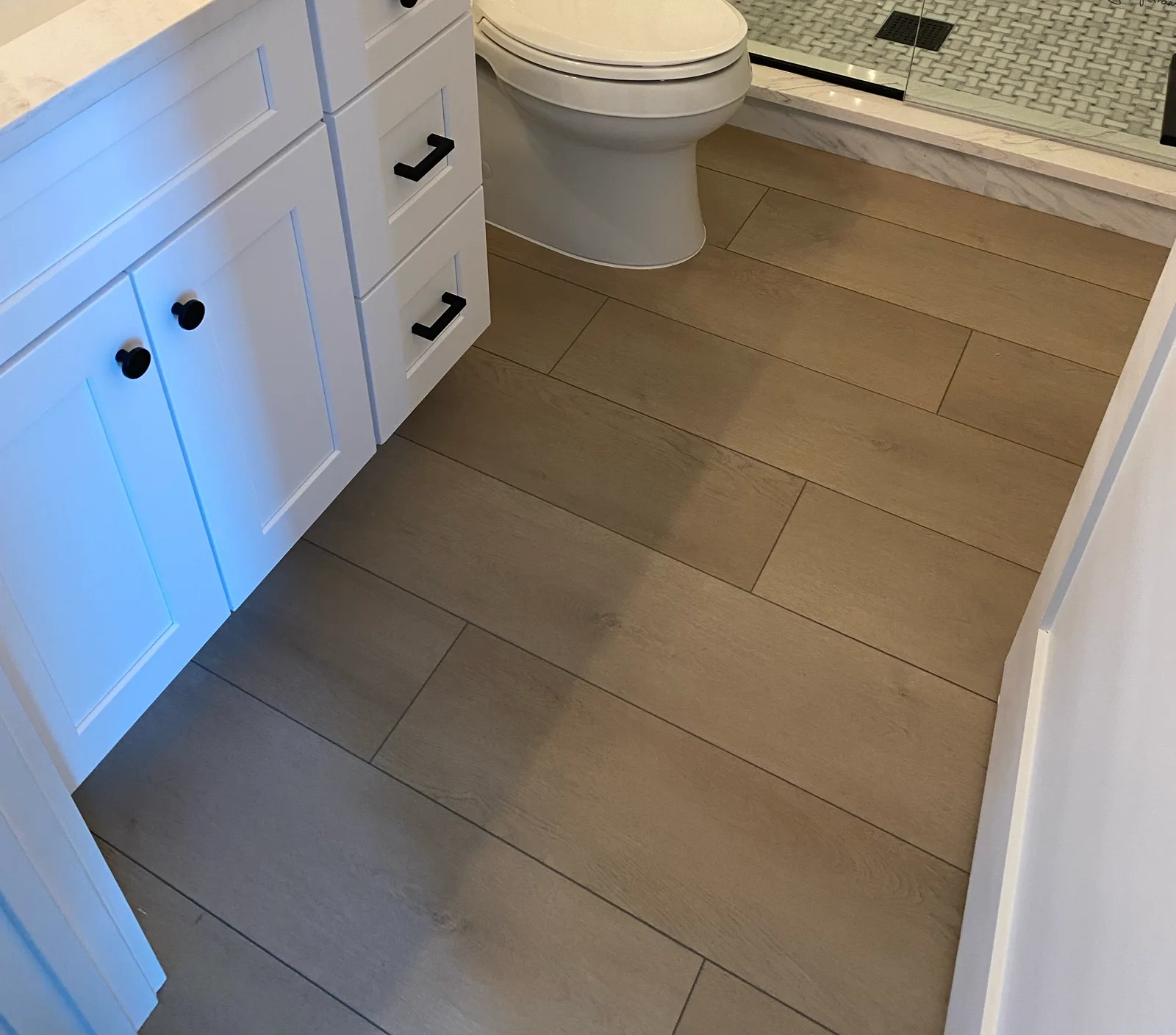 Style SPC Luxe 6.5mm greige wide-plank luxury vinyl flooring running through a remodeled bathroom past a white shaker vanity to a marble-look walk-in shower