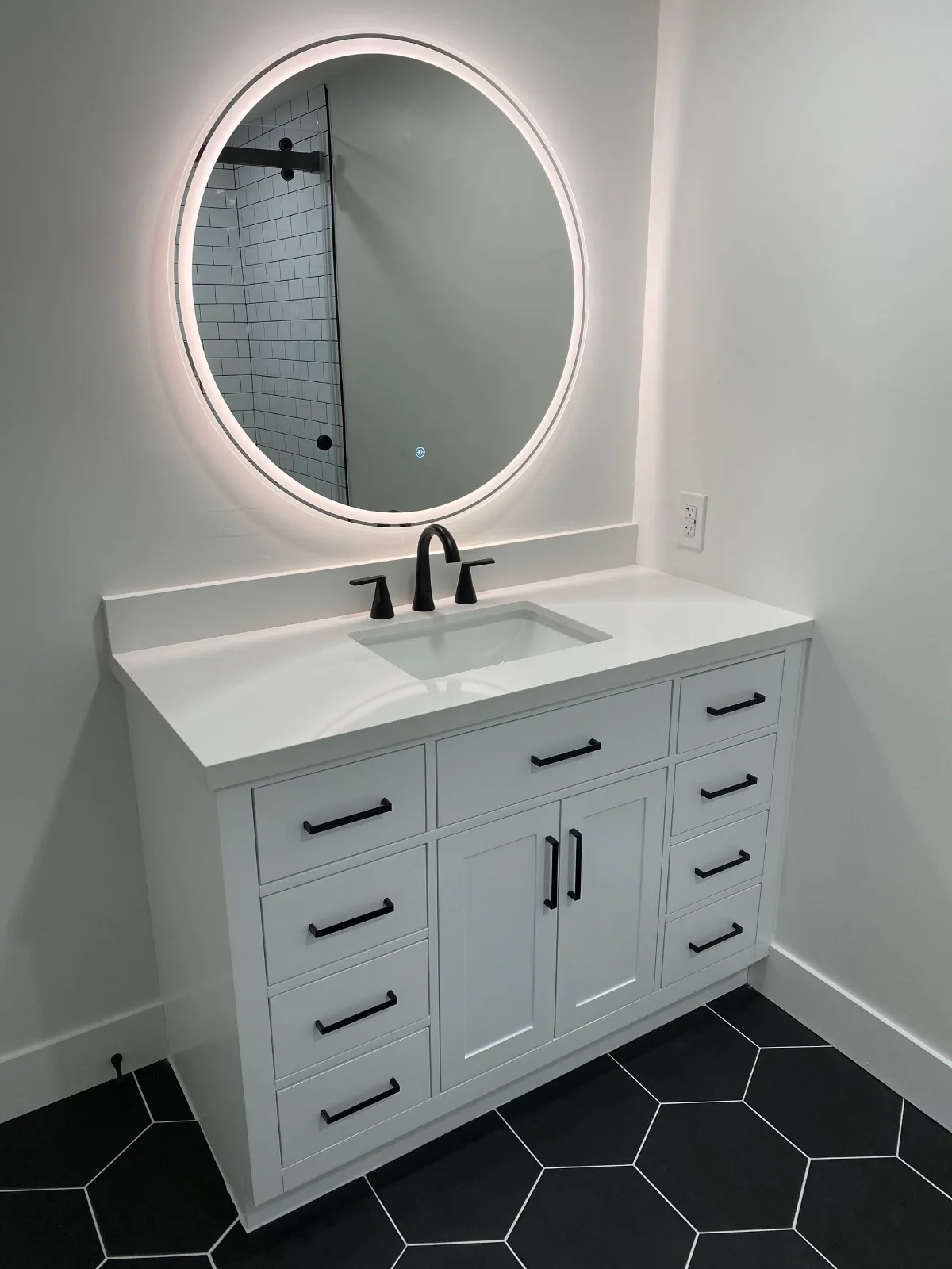 Round backlit mirror over a white vanity on black hexagon floor tile