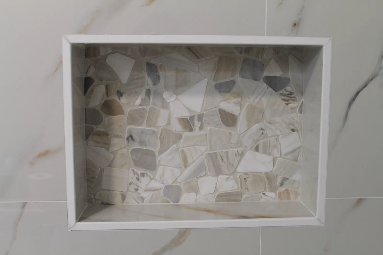 Recessed shower niche lined in natural pebble mosaic on calacatta-look tile