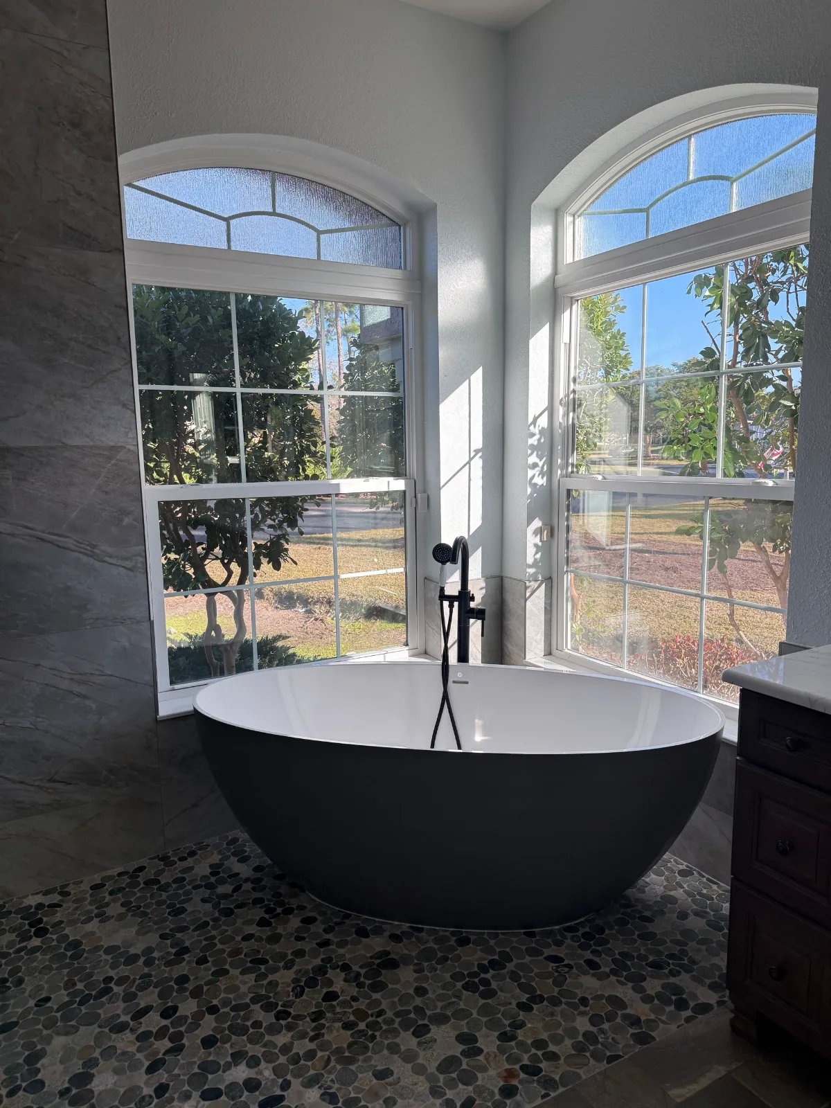 Matte black freestanding soaking tub on a river pebble floor beneath a window wall — Cape Coral master bath